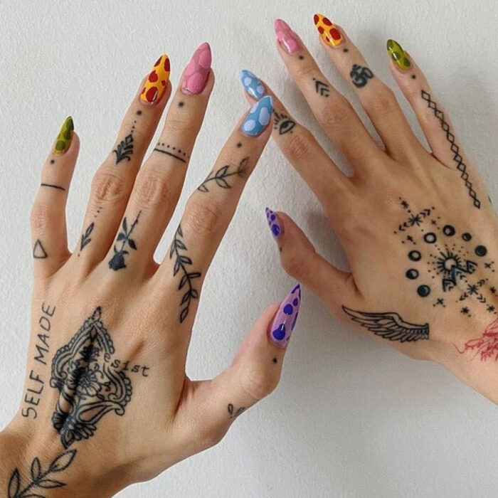 Manos cuidadas con manicura, uñas largas stiletto pintadas de colores verde, amarillo, rosa, azul y morado con lunares, además de dedos con varios tatuajes artísticos