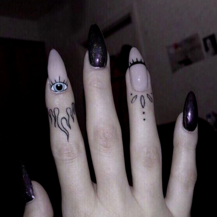 Manos elegantes con manicura, uñas largas stiletto pintadas en rosa pálido y negro con brillos, con un diseño de ojos y pestañas, y dedos tatuados en estilos tribales y con llamas