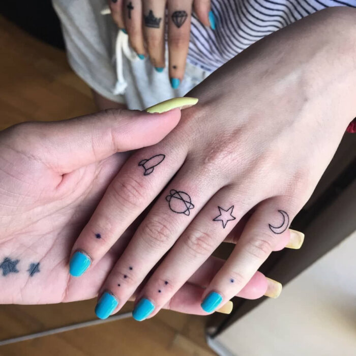 Manos cuidadas con manicura, uñas cortas y cuadradas pintadas de azul claro, dedos con tatuajes cósmicos de puntos, planetas, cohetes, luna, estrella y más