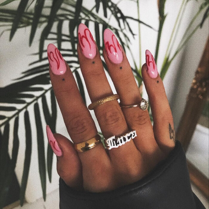 Manos cuidadas con manicura, uñas largas en forma de almendra con esmalte rosa y diseños de llamas rojas, dedos con tatuajes de puntos y rayos, y anillos dorados