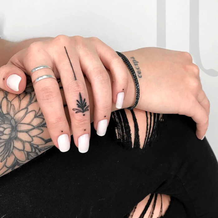 Manos elegantes con manicura, uñas cortas cuadradas pintadas de blanco, dedos tatuados de forma minimalista con plantas, puntos y líneas, usando anillos plateados