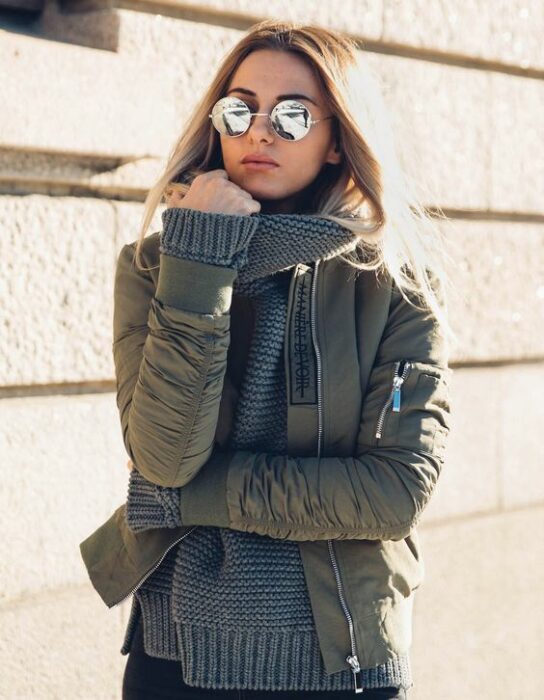 Chica con chamarra bomber verde militar, suéter gris y lentes de sol