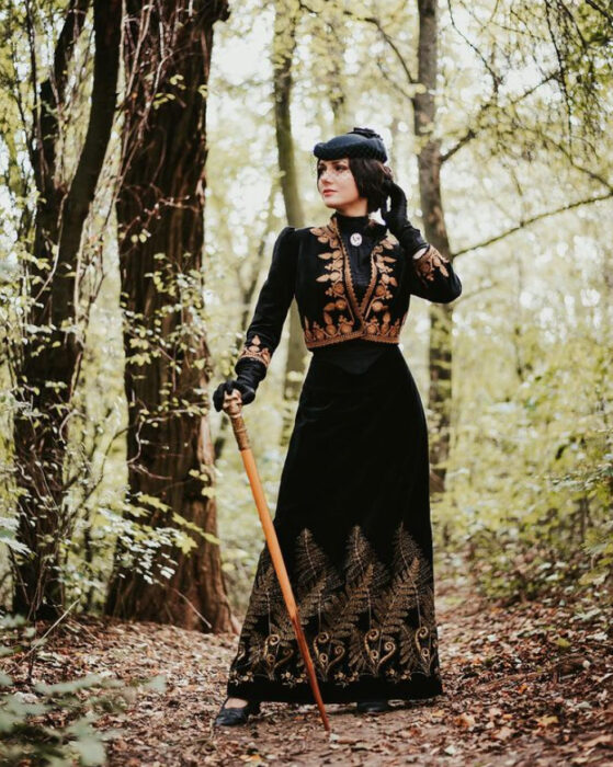 Mila Povoroznyuk usando atuendo de época, de falda y saco color negro con bordado dorado, tocado de sombrero y bordón