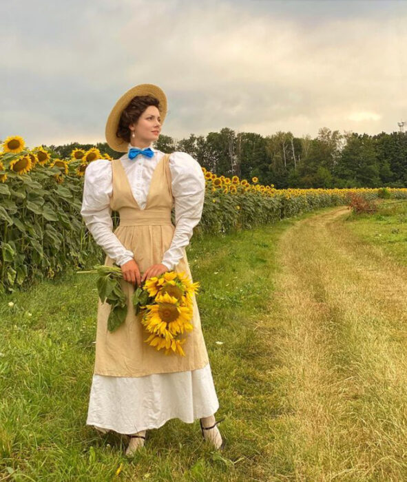 Mila Povoroznyuk usando vestido de época color blanco con ensamble color caqui y cargando unos girasoles