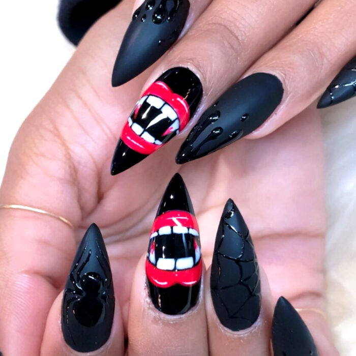 Manos con uñas largas stiletto pintadas en negro mate, con diseño de arañas, telarañas y labios rojos con colmillos de vampiro