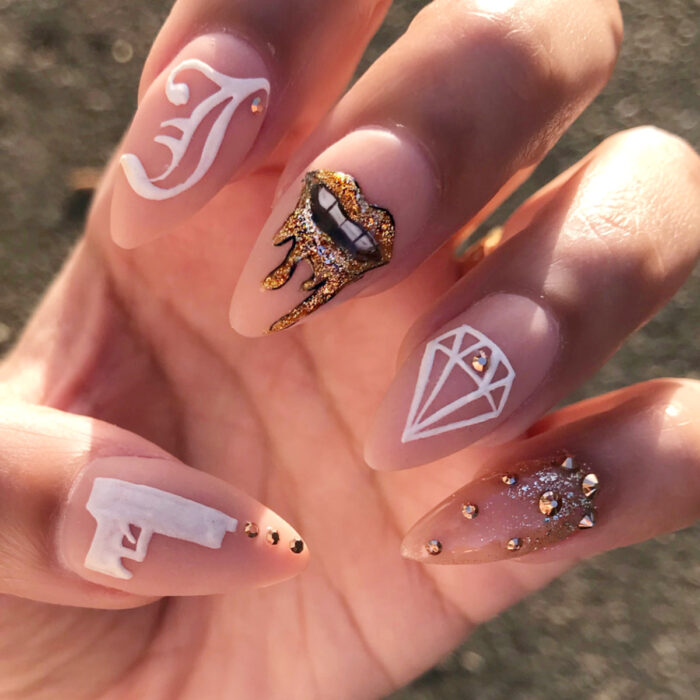 Manos con uñas largas en forma de almendra pintadas en esmalte nude, con diseño de labios y diamantina dorada