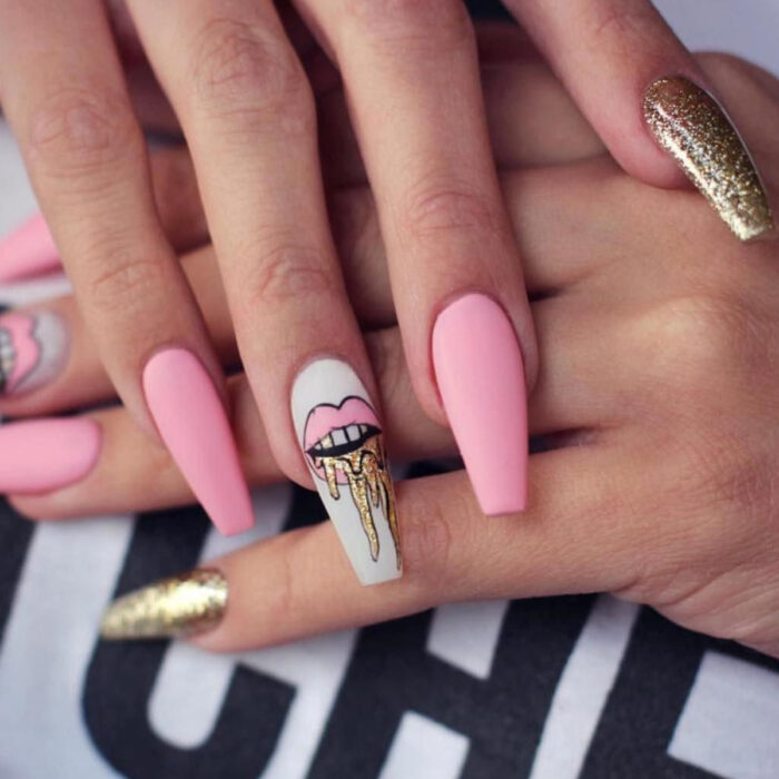 Manos de mujer con uñas largas cuadradas de bailarina, pintadas en esmalte mate rosa y blanco, con diseño de labios y brillo dorado en el meñique
