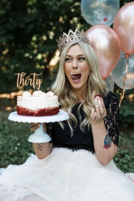 Chica rubia sosteniendo un pastel mientras usa una blusa negra y una diadema champagne