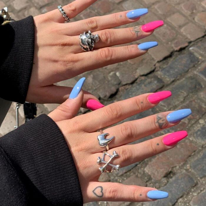 Diseños bonitos de manicura coloridos; manos de mujer con anillos rudos de calaveras, dientes y huesos, con tatuajes en los dedos de corazón, puntos y flores minimalistas, uñas largas cuadradas pintadas con esmaltes de colores azules y rosa