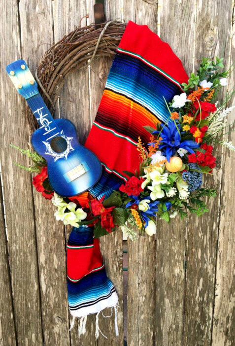 Decoración de Navidad con temática mexicana; corona para la puerta con ramas, rebozo de colores rojo, azul y anaranjado, guitarra pequeña y flores de plástico