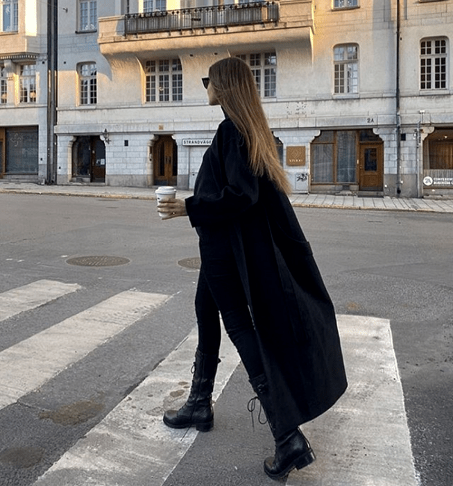 chica rubia usando lentes de sol, abrigo largo negro, skinny jeans negros, botines negros de plataforma y top negro de cuello alto