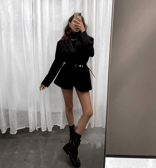chica de cabello castaño usando un top de cuello alto negro, blazer negro, cinturón negro de cuero, shorts de ciclismo negros, botas de cuero negras con plataforma y suela gruesa