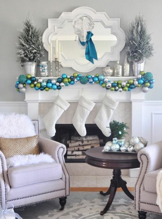 Sala blanca con chimenea decorada con esferas turquesas, plateadas y verdes, además de tres botas blancas colgantes
