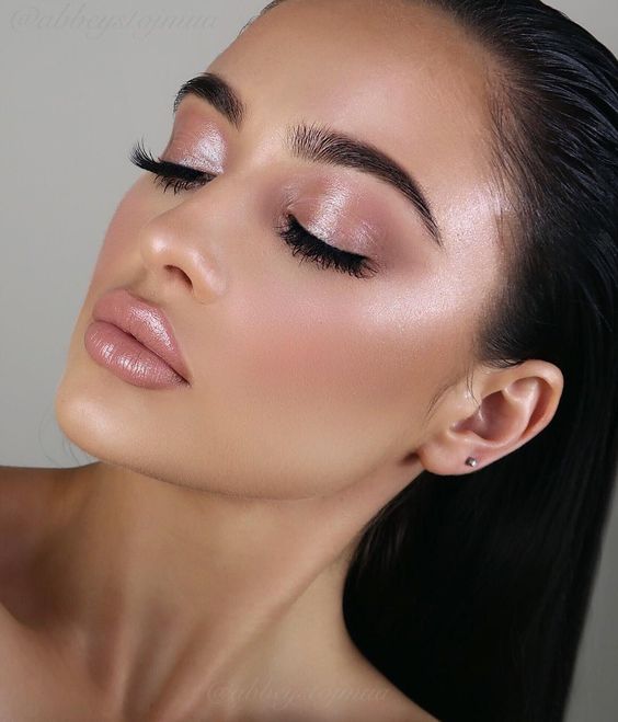 Mujer morena clara con coleta cierra los ojos y usa maquillaje natural en color rosita con un toque de brillo y un labial rosa con gloss