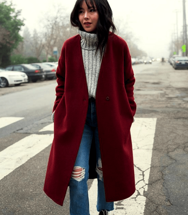 mujer de cabello castaño con suéter gris, abrigo rojo y jeans ajustados rotos