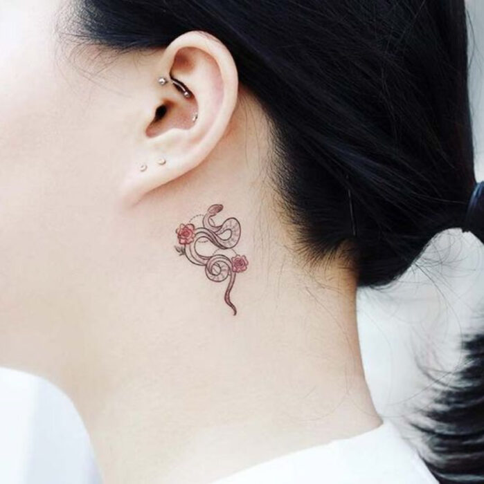 Tatuaje pequeño de una serpiente en tinta negra con flores rojas en la zona detrás de la oreja