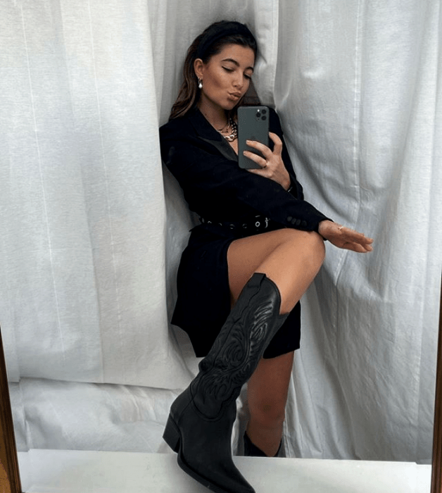 mujer rubia usando un blazer oversized negro como vestido, cinturón con estoperoles, botas negras de cuero estilo vaquero y diadema negra