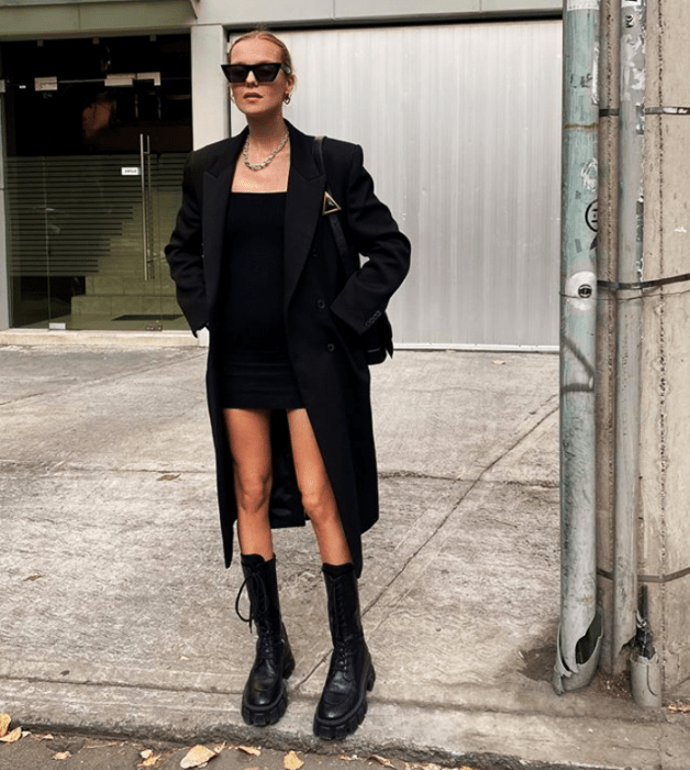 mujer rubia usando lentes de sol, collar de cadena, vestido mini negro, abrigo largo negro, botas de cuero negras y bolso negro mini