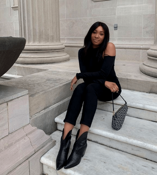 mujer morena usando un top de manga larga negro, skinny jeans negros, bolso de mano negro y blanco, y botines negros