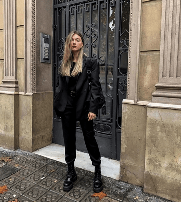 mujer rubia usando un top negro, saco negro, pantalones de vestir negros, botas de cuero negras y un bolso pequeño negro
