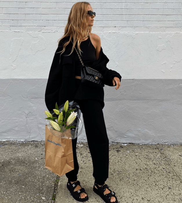 mujer rubia usando lentes de sol, top sin mangas negro, sudadera negra, sandalias negras y un bolso pequeño negro