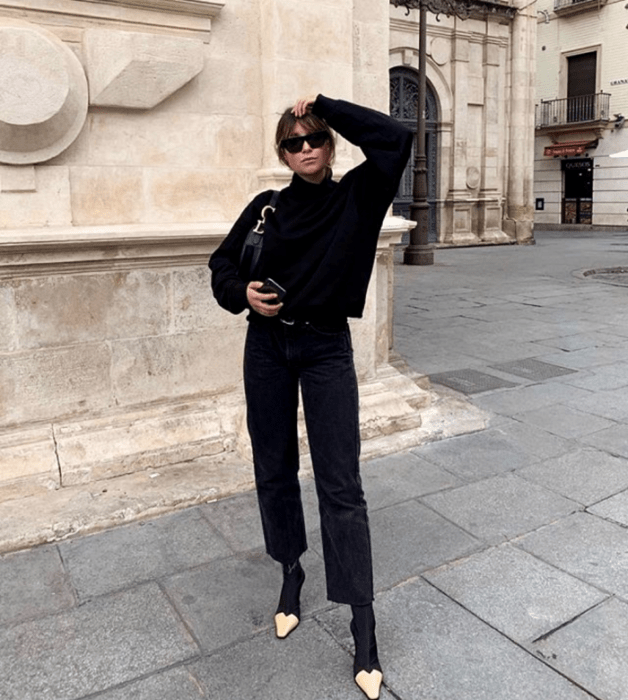 mujer de cabello castaño usando lentes de sol, suéter negro de manga larga, jeans rectos negros, botines negros con punta beige y un bolso pequeño de mano