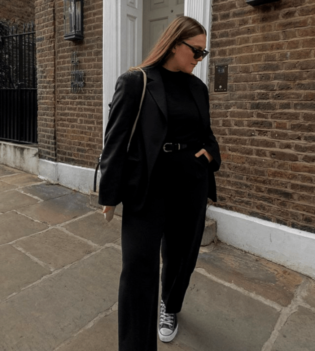 mujer rubia usando lentes de sol, top negro, cinturón de piel, pantalones de vestir negros, converse negros y saco negro