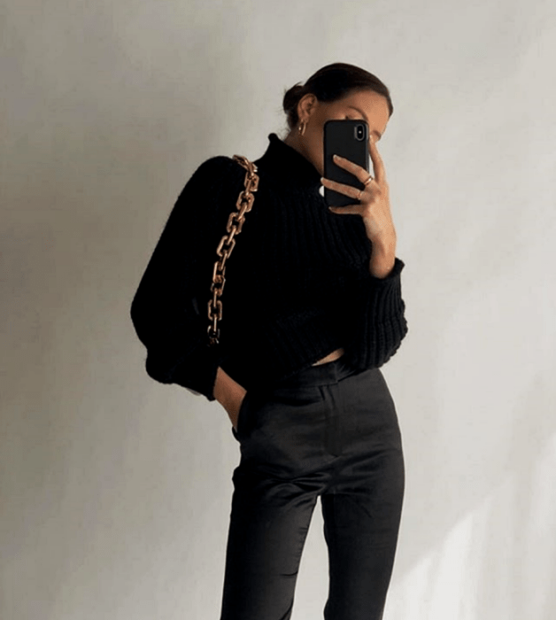 mujer de cabello castaño usando un suéter de cuello alto negro, pantalones de vestir negros y un bolso con tirante de cadena dorado