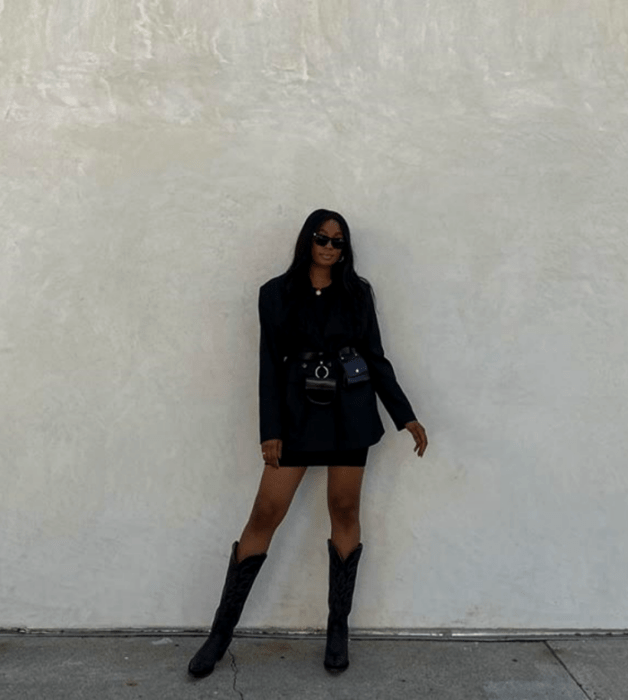 mujer morena usando lentes de sol, saco oversized negro, falda negra, botas largas de cuero estilo vaquero y un bolso de cuero negro a la cintura