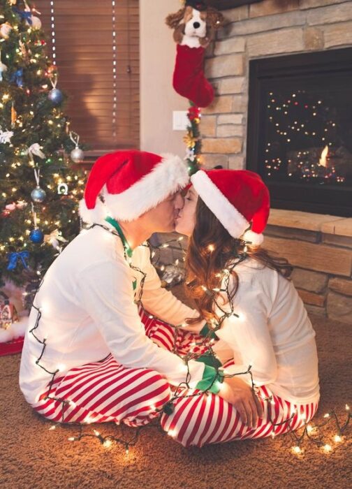 Pareja sentada de frente con el árbol de navidad detrás de ellos, besándose y rodeados de luces navideñas