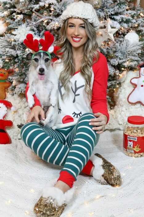 Chica usando pijama de navidad, sentada delante del árbol navideño, acompañada de su perro con una diadema de reno