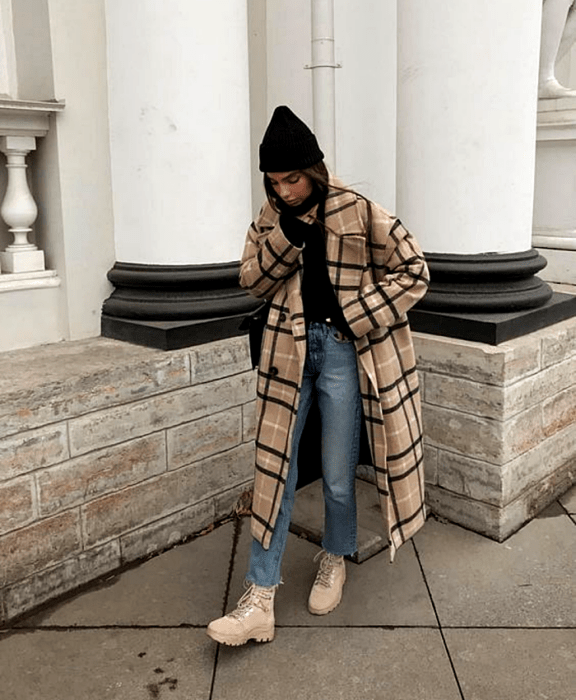 chica de cabello castaño usando un gorro beanie negro, abrigo largo café con líneas blancas y negras, jeans a la cintura, top negro y botas cafés de montaña