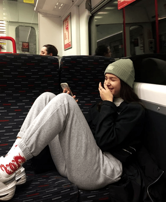 chica de cabello negro usando un beanie gorro verde, sudadera negra, pantalones deportivos grises, calcetas blancas con dragón rojo y tenis blancos