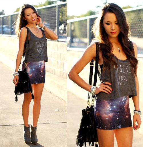 Chica morena, delgada y con cabello largo ondulado posando en la calle con blusa de tirantes gruesos color gris, falda con estampado de galaxia, botines negros y bolsa negra colgando de su hombro derecho