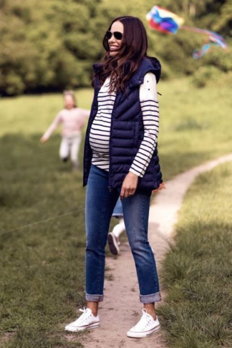 Chica usando un outfit de maternidad de jeans, tenis, chaleco y blusa de rayas
