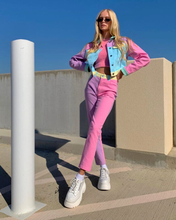 Look colorido aesthetic; mujer rubia en estacionamiento con outfit colorido, cabello lacio y largo, crop top rosa, jeans en tonos pastel, chamarra a juego, tenis chunky blancos, lentes de sol cuadrados