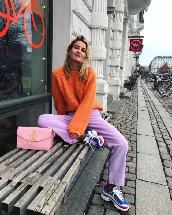 Look colorido aesthetic; mujer sonriendo sentada en banca de madera, cabello rubio lacio y despeinado, atuendo colorido, suéter tejido anaranjado, pants morado lila, tenis chunky azules y rosa mexicano, bolsa pastel con cadena dorada