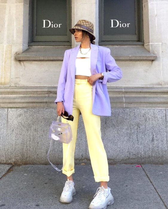 Look colorido aesthetic; mujer castaña con gorro de pescador y atuendo colorido, crop top blanco, pantalón amarillo pastel, blazer morado, bolsa transparente, tenis blancos, frente a tienda Dior