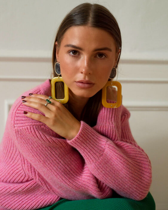 Look colorido aesthetic; mujer de ojos cafés, cabello lacio suelto, aretes amarillos retro, suéter rosa, falda y anillos verdes, uñas rojas