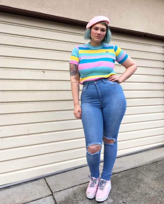 Look colorido aesthetic; mujer plus size con la mano en la cintura, atuendo colorido, boina rosa pastel, blusa de manga corta azul fuerte y clara, jeans desgastados, tenis en tonos pastel, cabello corto con tinte fantasía, tatuaje de rosas