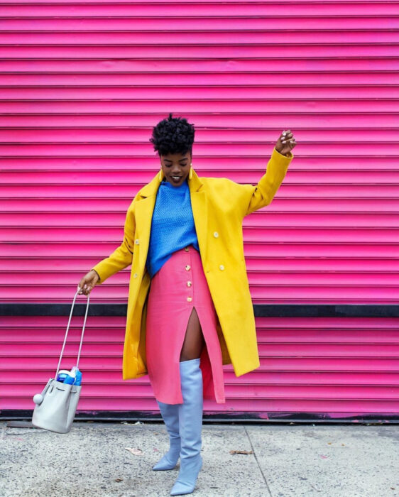 Look colorido aesthetic; mujer afroamericana con cabello chino, sonriendo, vistiendo colores, blusa azul, falda rosa con abertura, botas largas grises, abrigo amarillo, bolsa y aretes grandes