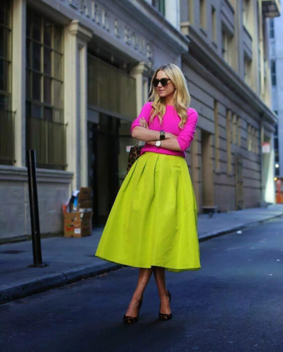 Look colorido aesthetic; chica posando en la calle, cabello rubio largo y ondulado, lentes oscuros grandes, suéter rosa mexicano, falda verde neón, zapatillas animal print