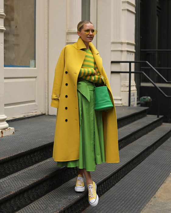 Look colorido aesthetic; mujer posando en escaleras, cabello rubio en coleta, lentes amarillos, gabardina, blusa a rayas amarillas y verdes, falda moderna, tenis converse
