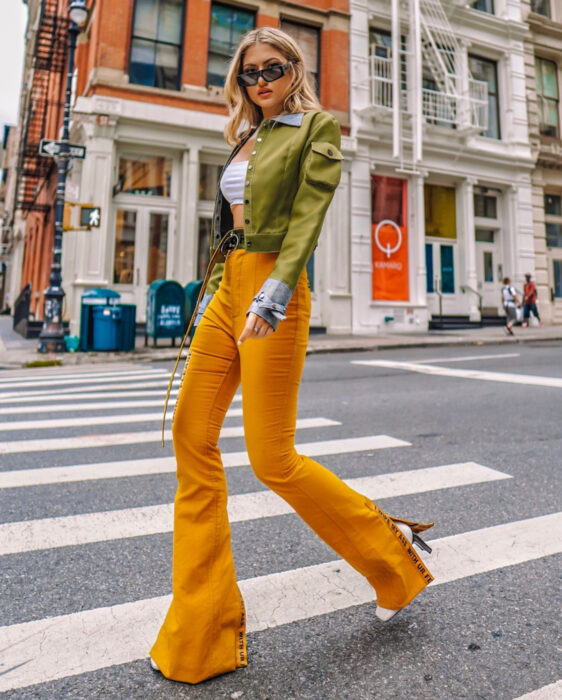 Look colorido aesthetic; mujer caminando en la calle, cabello rubio, mediano y ondulado, lentes rectangulares, crop top blanco, chamarra verde militar, pantalones acampanados naranja, botines blancos
