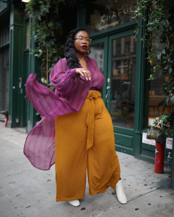 Look colorido aesthetic; mujer afroamericana con cabello negro trenzado, lentes retro, labial vino, blusa de gasa rosa con rayas verticales, pantalón ligero mostaza, botines blancos