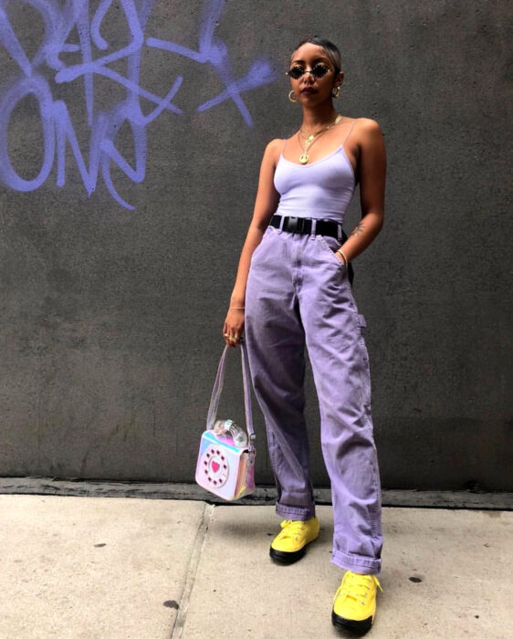 Look colorido aesthetic; mujer afroamericana con cabello corto, lentes pequeños, blusa lila, mom jeans, tenis amarillos, bolso en forma de teléfono antiguo holográfico