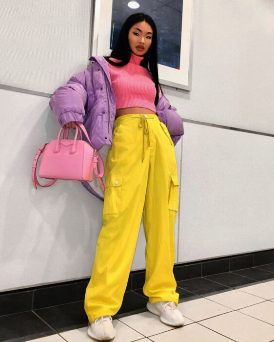Look colorido aesthetic; mujer de ojos rasgados, cabello negro lacio, recargada en la pared, atuendo colorido, crop top rosa, chamarra morada, pantalones holgados amarillos, tenis blancos, bolsa de mano