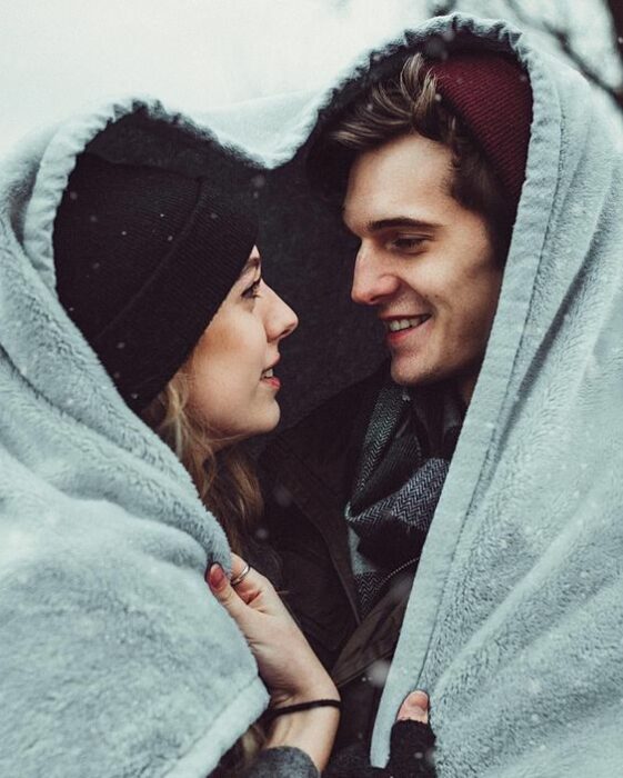 Mujer rubia con gorrito negro y hombre de cabello castaño con gorrito guinda se cubren con una manta color azul formando un corazón