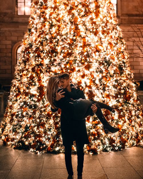 Chico alzando a chica rubia de cabello largo frente a árbol de navidad gigantes con luces encendidas