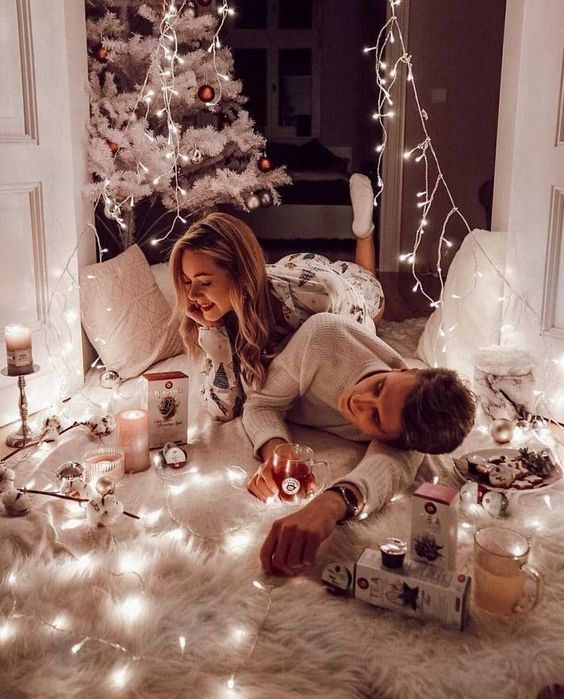 Pareja con pijamas blancas acostados y riendo en una alfombra blanca con luces navideñas en el piso y un árbol de navidad en el fondo